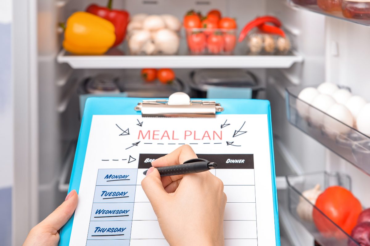 Woman looking at open fridge writing on meal plan