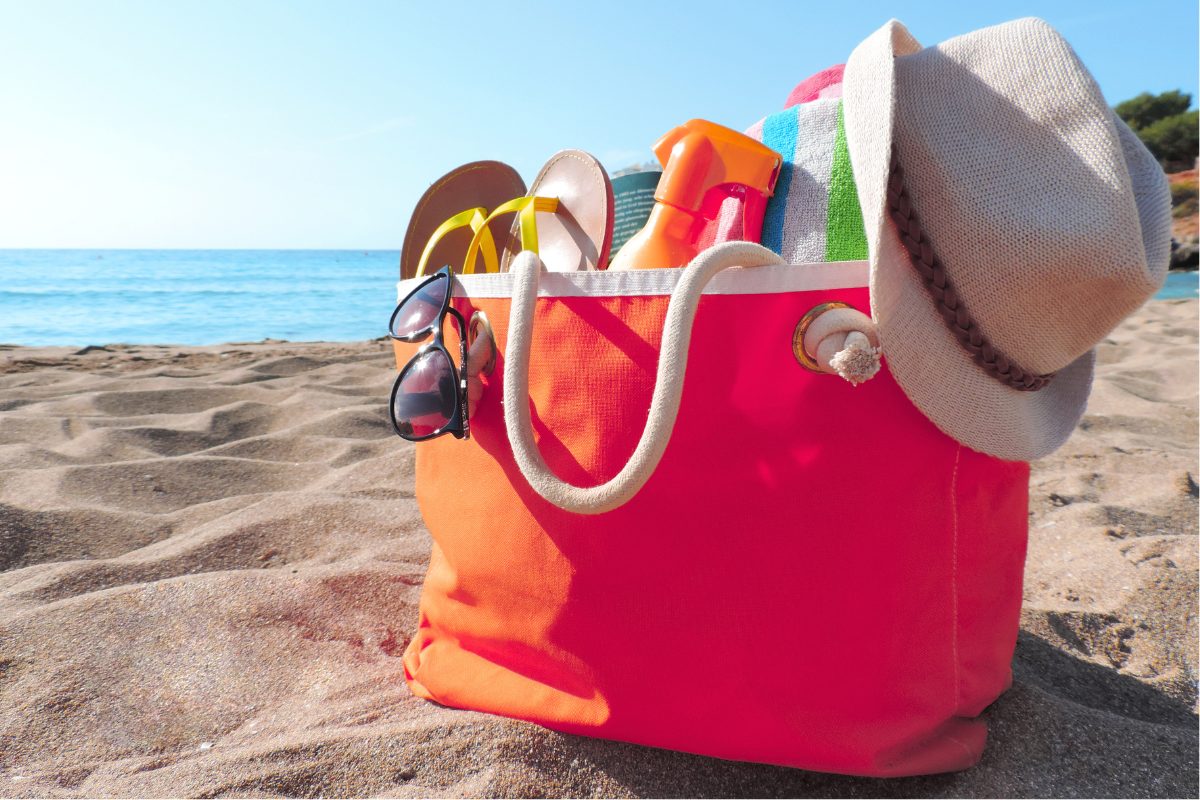 A bag sitting on the sandy beach near the water, filled with beach toys, hats and other summer items.